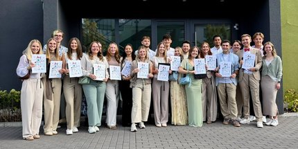 Absolventinnen und Absolventen des Kurses Ph24 der Physiotherapieschule Rhein-Nahe stehen vor dem Gebäude der Schule und halten Ihre Abschlussurkunden in die Kamera.