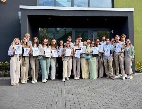 Absolventinnen und Absolventen des Kurses Ph24 der Physiotherapieschule Rhein-Nahe stehen vor dem Gebäude der Schule und halten Ihre Abschlussurkunden in die Kamera.